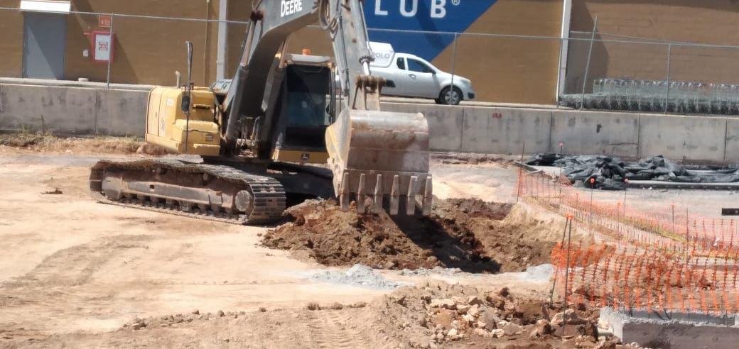 Construction site with a small yellow excavator digging into the dirt, surrounded by orange safety fencing and construction materials.