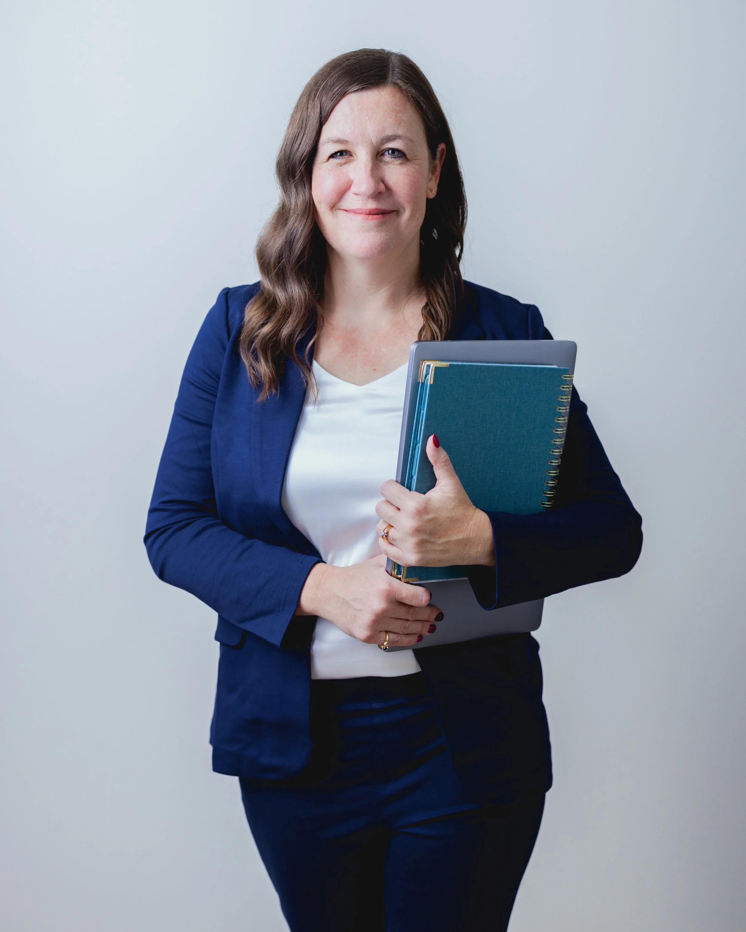 A woman with long brown hair wearing a navy blazer and white top holding a blue notebook and folder, smiling at the camera, against a plain light-colored background.