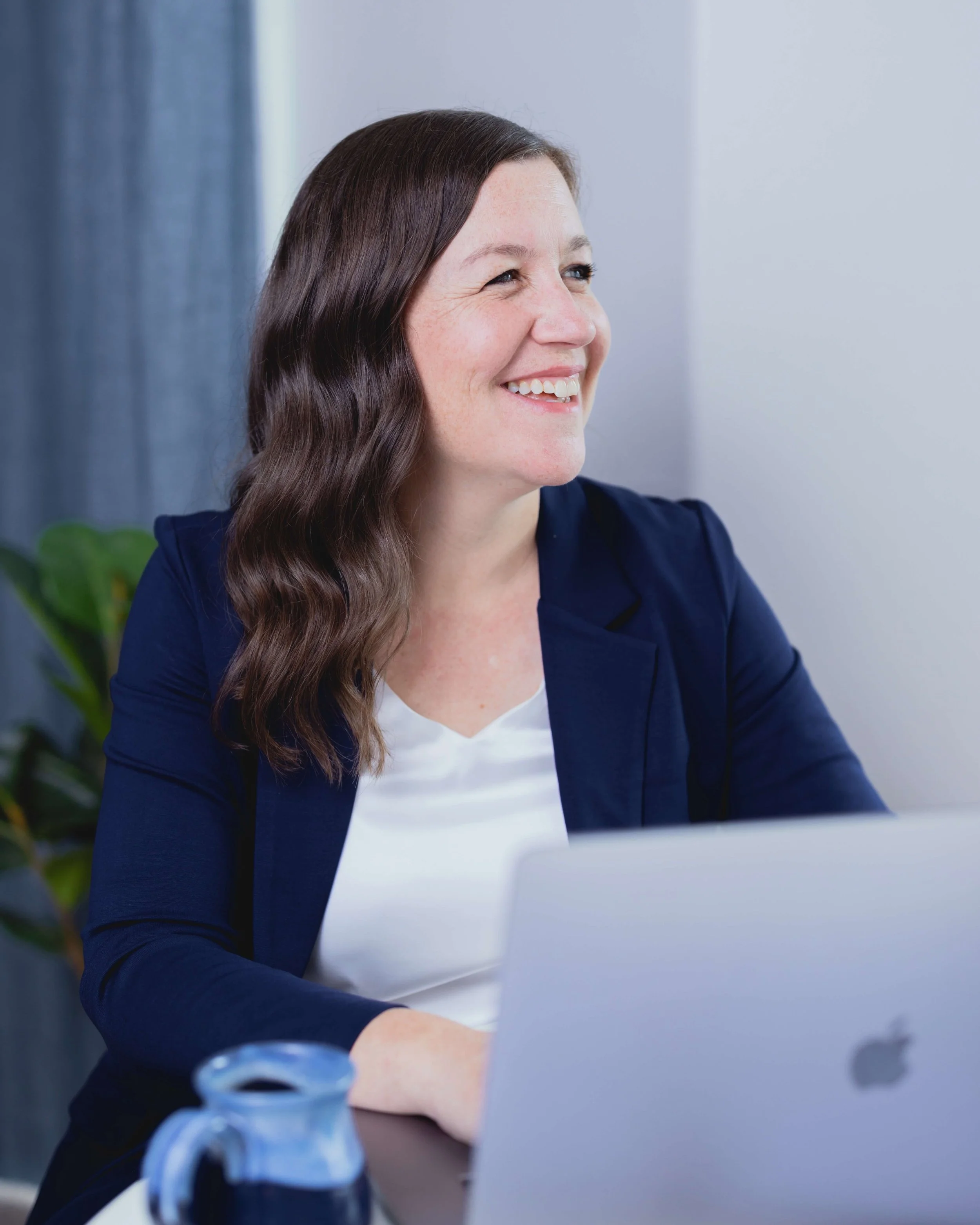 A woman smiling with long brown hair, wearing a navy blazer, sitting at a desk with a laptop, in front of a light-colored wall with a green plant nearby.