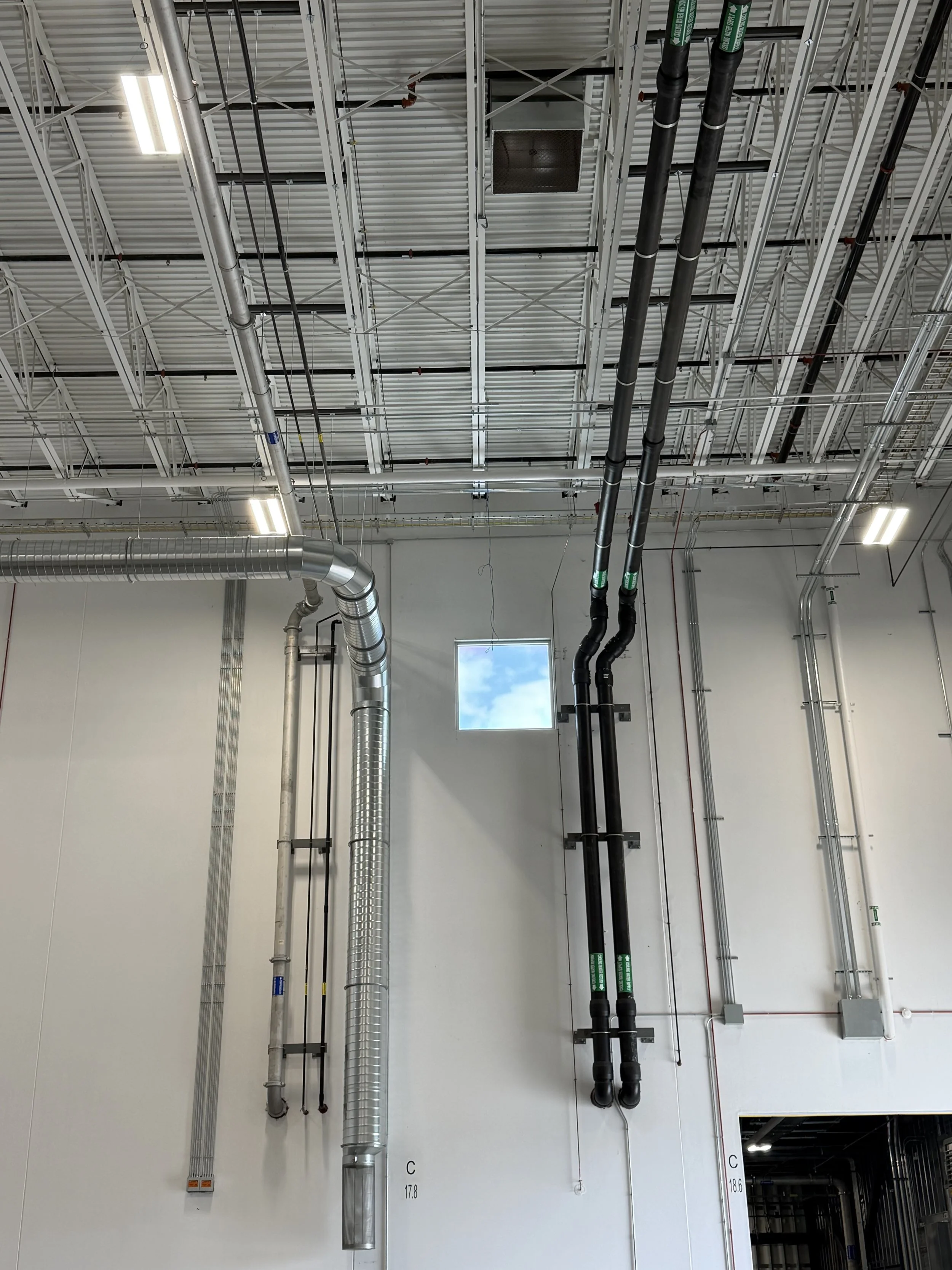 Industrial ceiling with metal framework, black and silver pipes, ductwork, and fluorescent lighting, with a small window showing the sky.