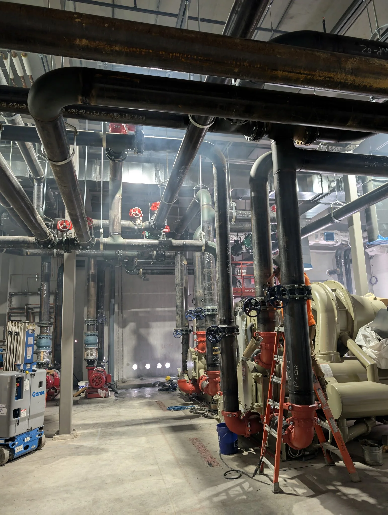 Industrial room with interconnected pipes, valves, and machinery, including a ladder and some equipment in the background.