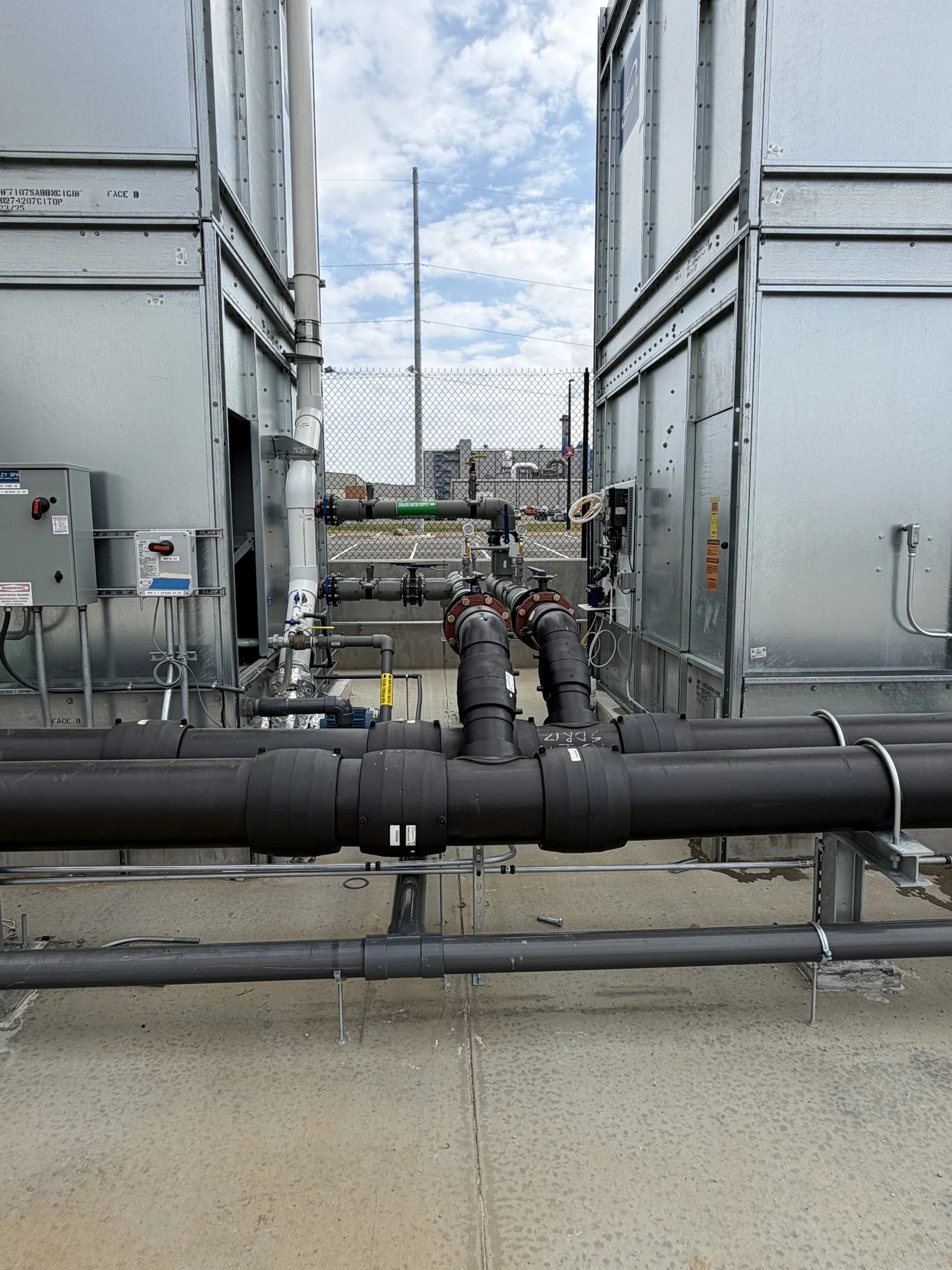 View of industrial HVAC or plumbing equipment with large black pipes, metal panels, and control boxes, set against a concrete ground and fenced outdoor background with blue sky and clouds.