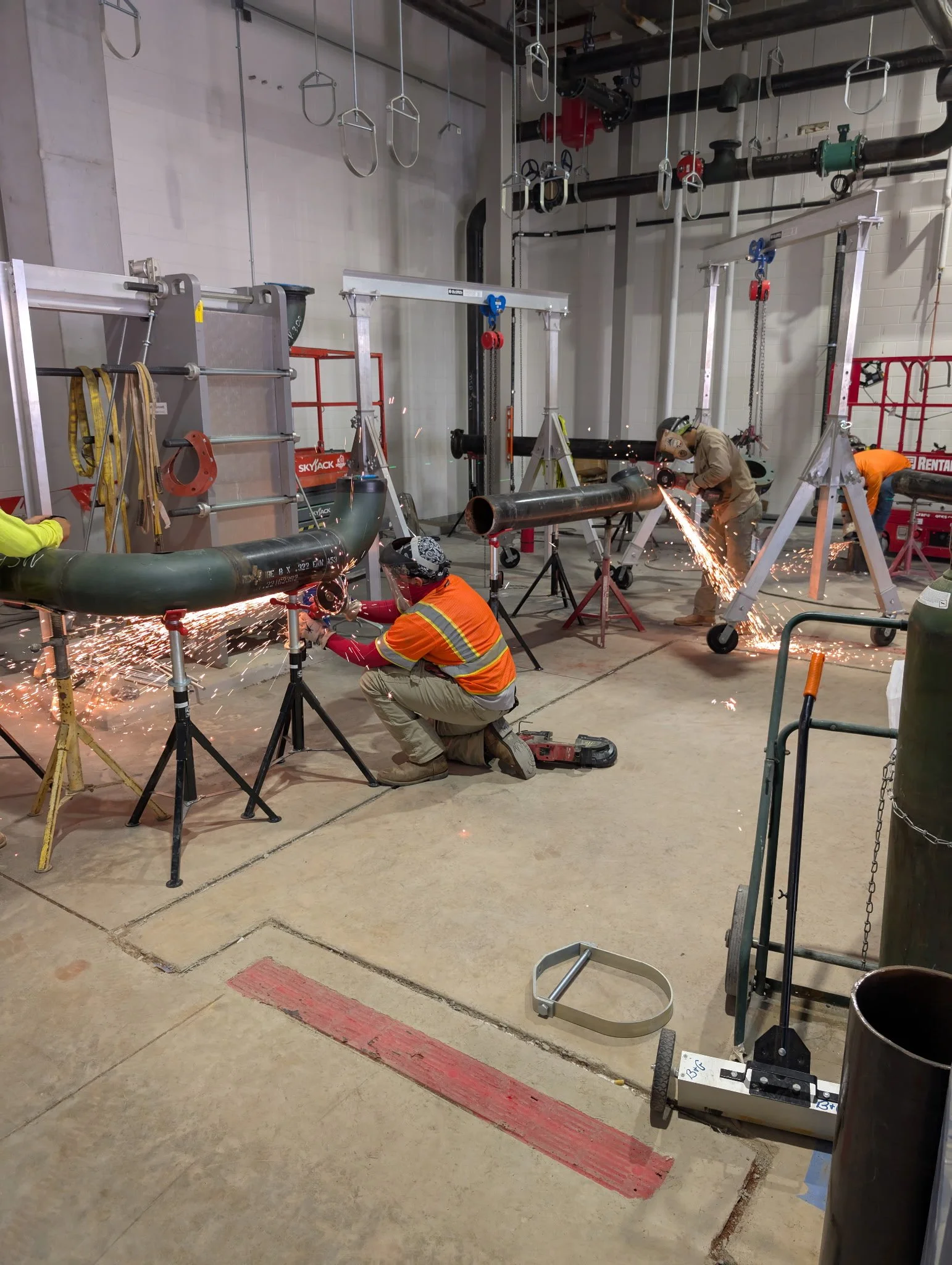 Workers are welding large pipes in an industrial setting, with sparks flying. Various tools, scaffolds, and industrial equipment are visible around them.