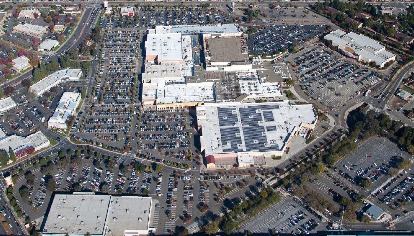 Aerial view of a shopping mall with multiple parking lots, surrounded by roads, commercial buildings, and some trees.