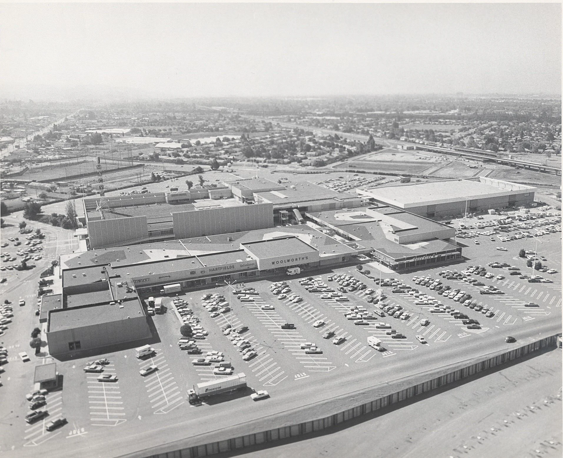 Aerial black and white photo of a shopping mall parking lot with many cars and a large retail store building, with surrounding roads and distant urban area.