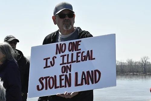 Man at a protest holding a sign that says "No one is illegal on stolen land"