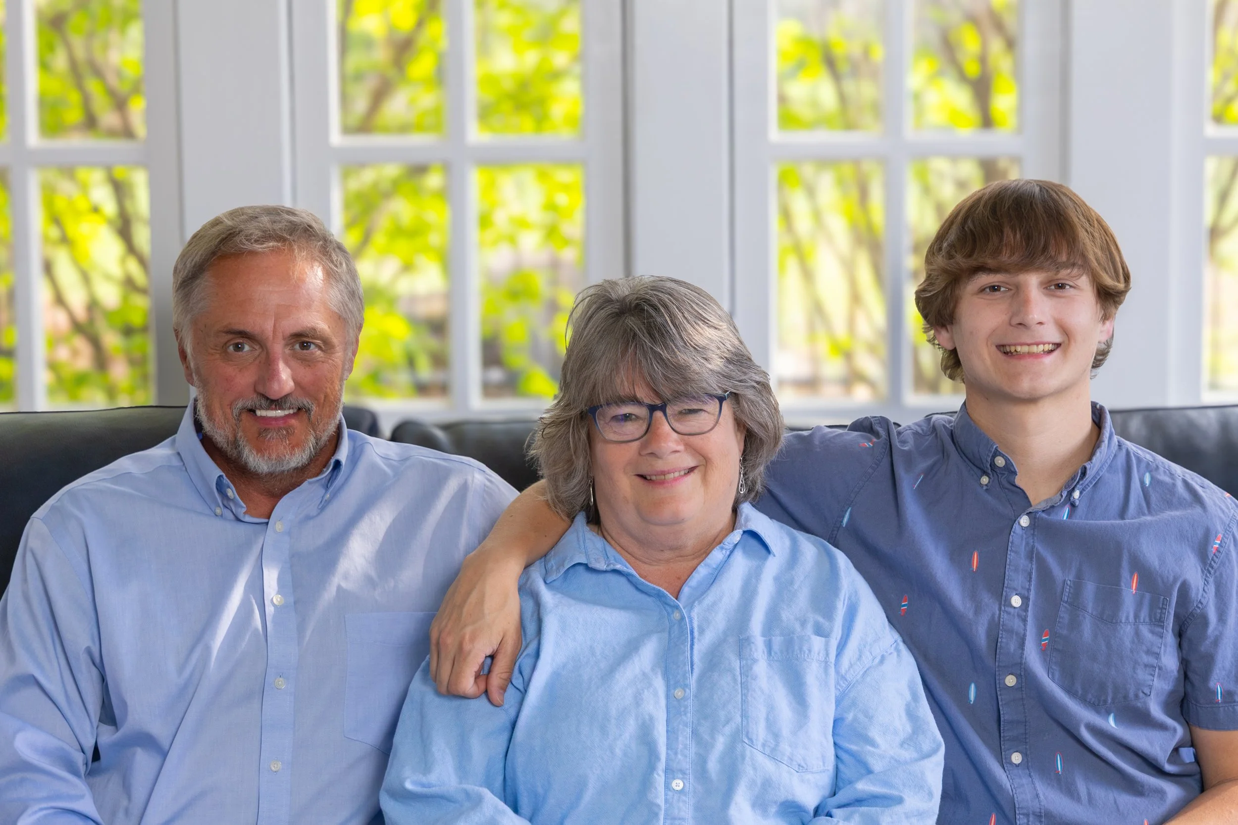 Three people sitting on a black couch in front of a window with green trees outside. The person on the left is an older man with gray hair, beard, and mustache, wearing a light blue shirt. The person in the middle is an older woman with gray hair and glasses, wearing a blue shirt. The person on the right is a young man with medium-length brown hair, smiling, wearing a blue collared shirt.