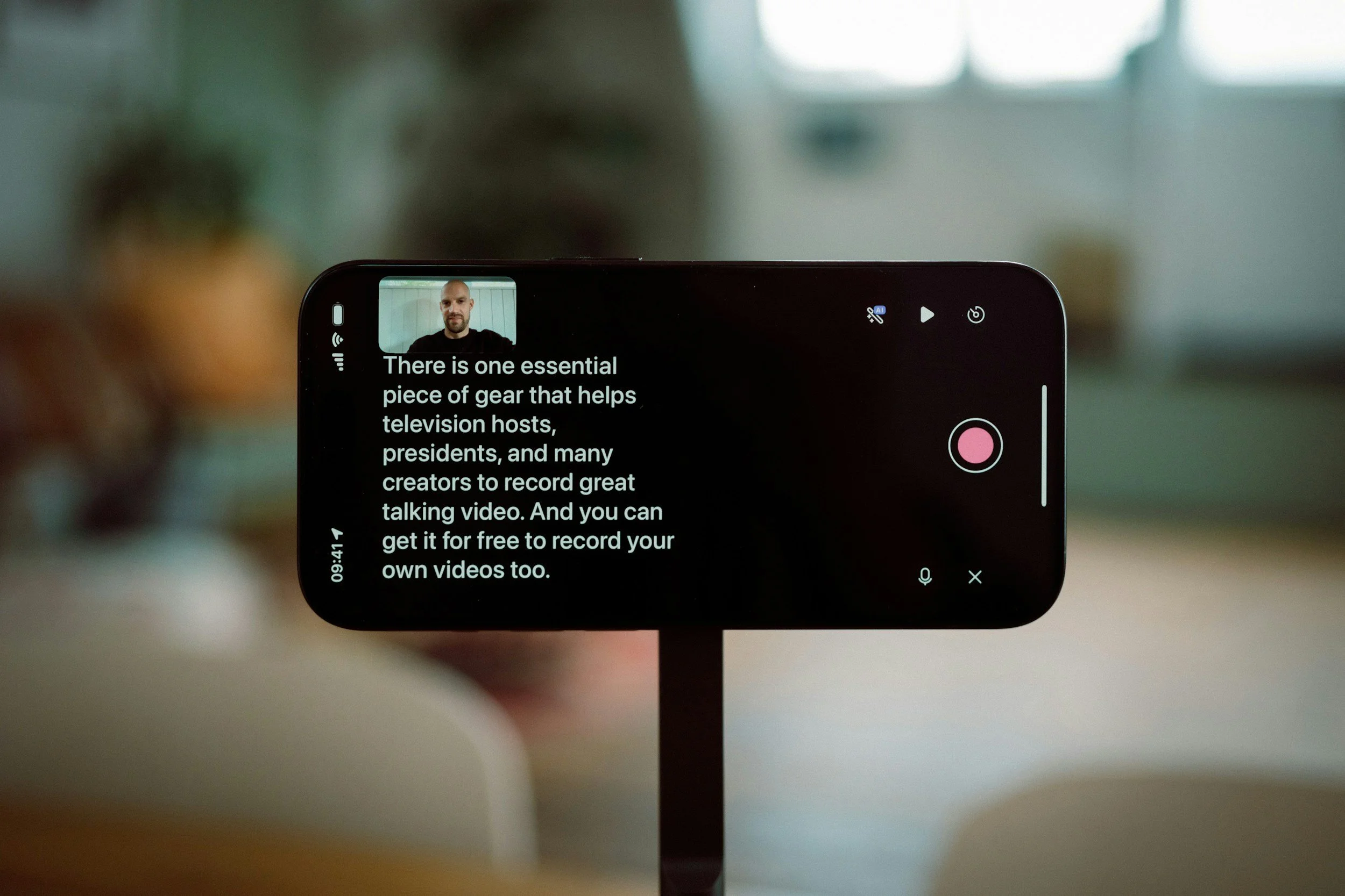 A smartphone on a stand shows a video recording app with a person speaking at the top left corner and a description text about gear for recording videos. The screen displays a pink record button, a microphone icon, and other control options, with a blurred background.