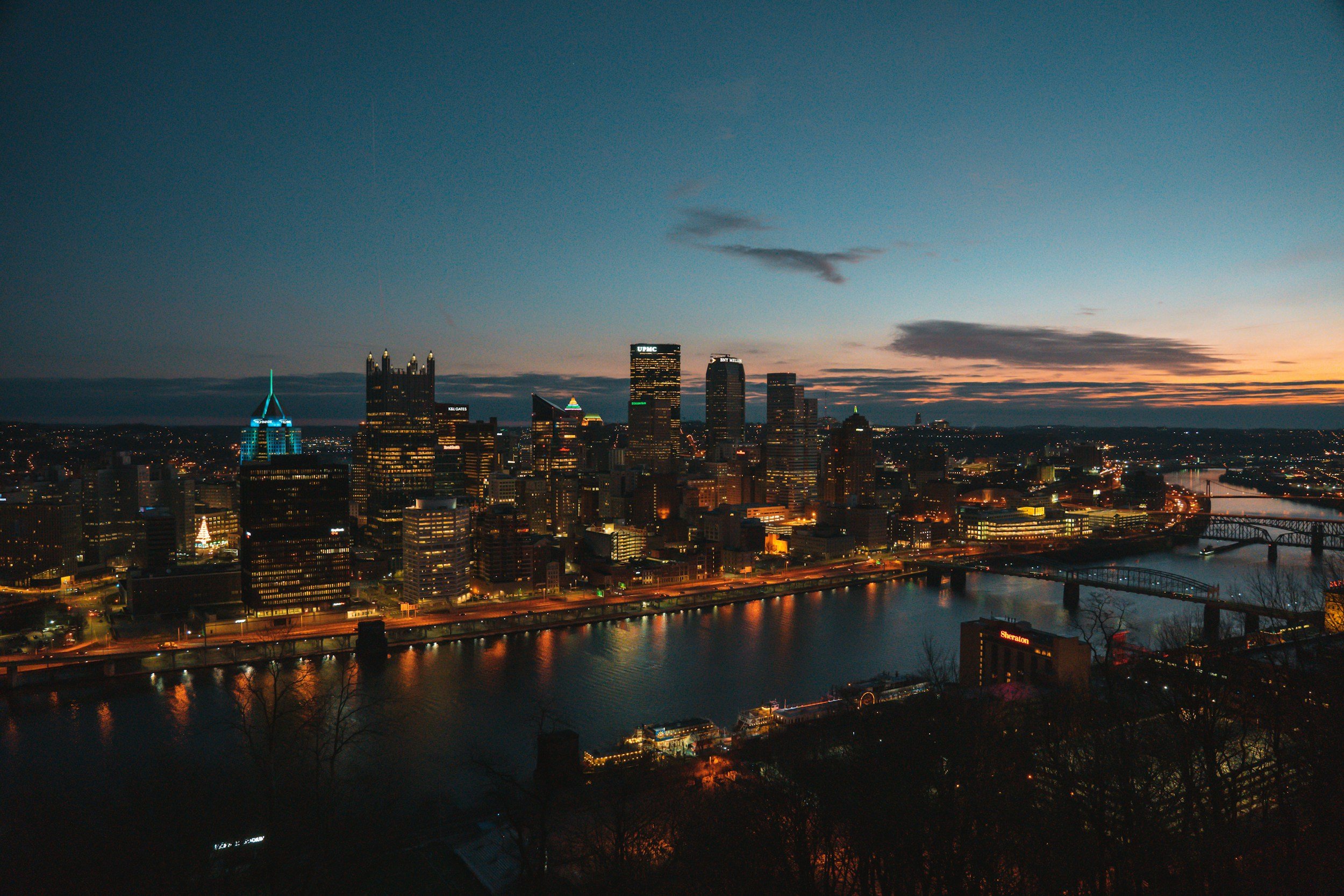 Nighttime view of Pittsburgh skyline with illuminated buildings reflecting on the Ohio River.
