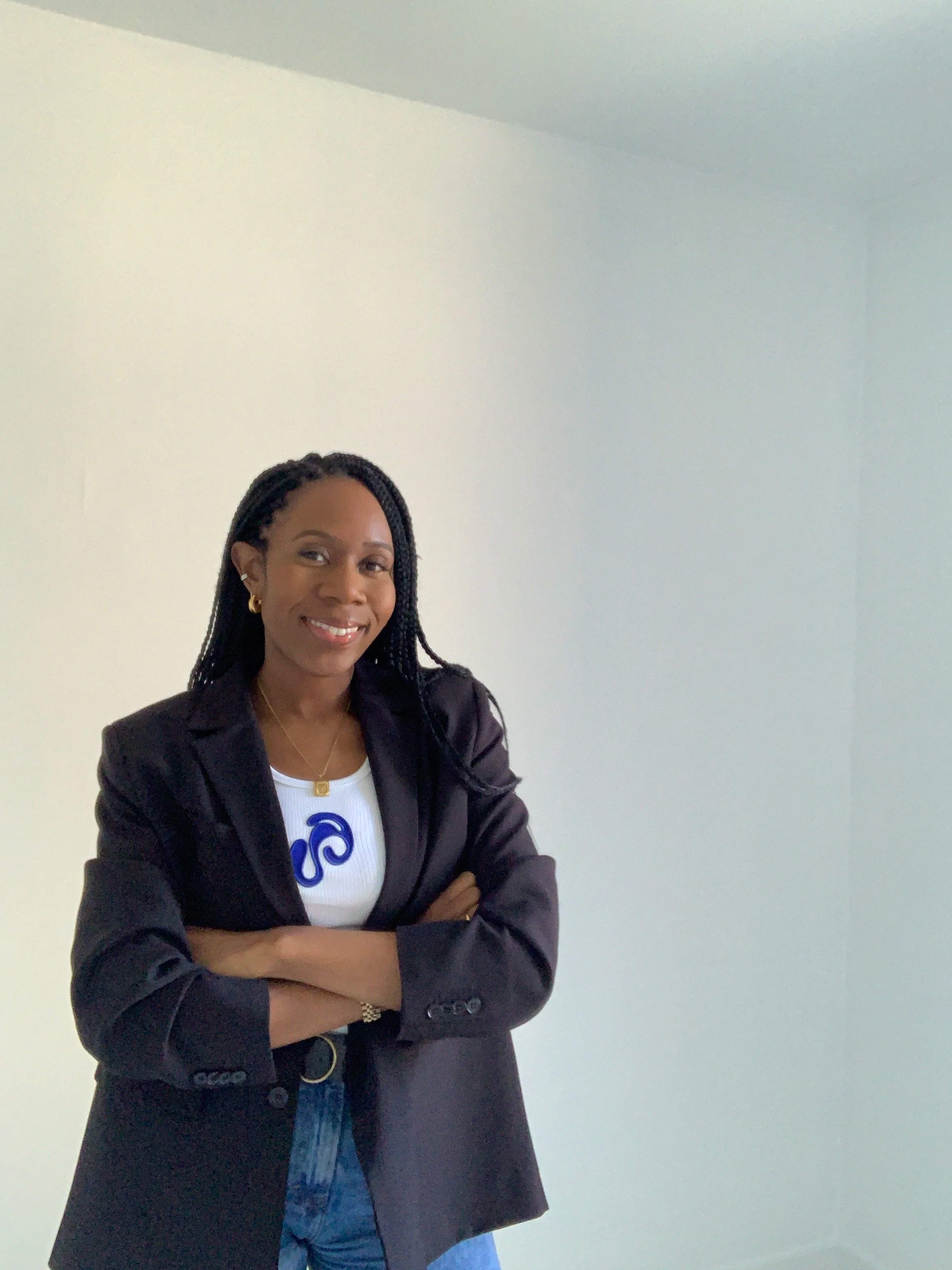 A woman with braided hair, wearing a black blazer over a white company logo t-shirt, standing with arms crossed in a plain white room, smiling at the camera.