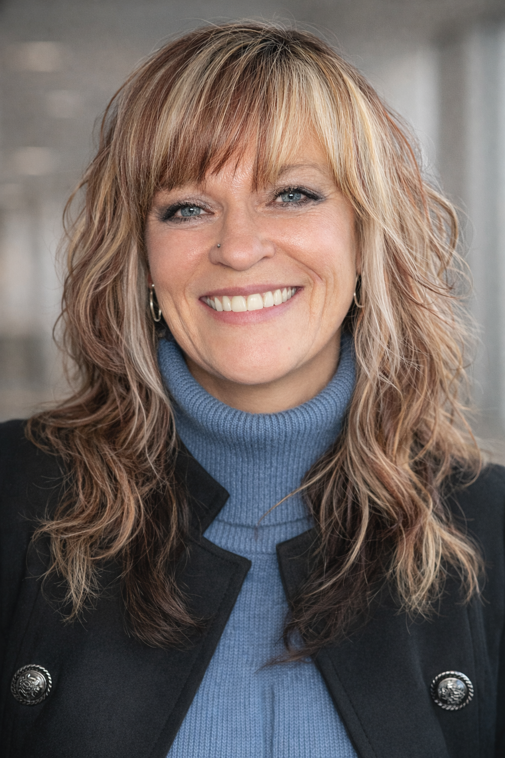 A smiling woman with shoulder-length wavy blonde hair, wearing a blue turtleneck sweater and a black jacket with decorative buttons, standing indoors.