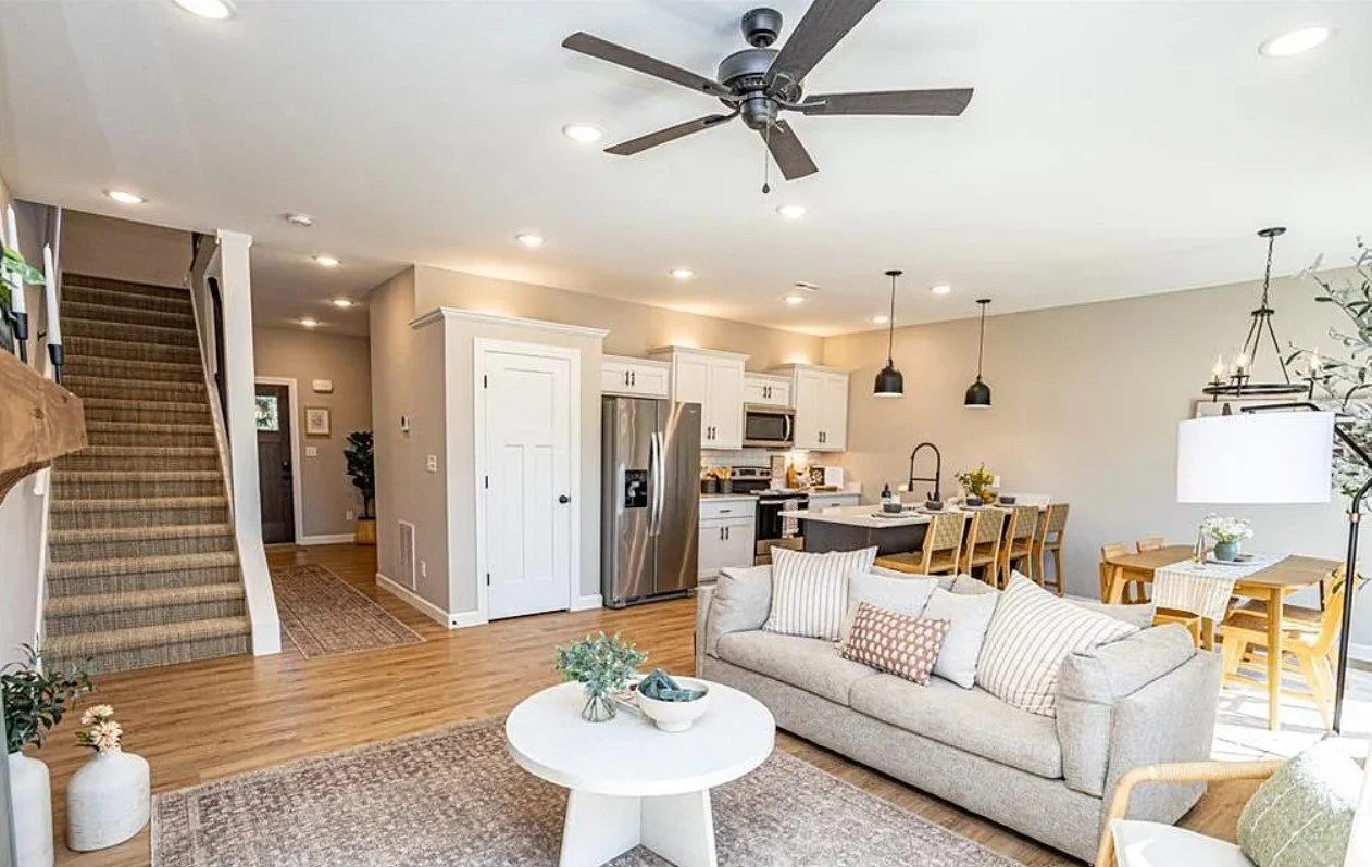Open-concept living room and kitchen with a ceiling fan, hardwood floors, a white sofa with pillows, a round coffee table with decorative items, a dining table with chairs, pendant lights, and a staircase with carpeted steps.