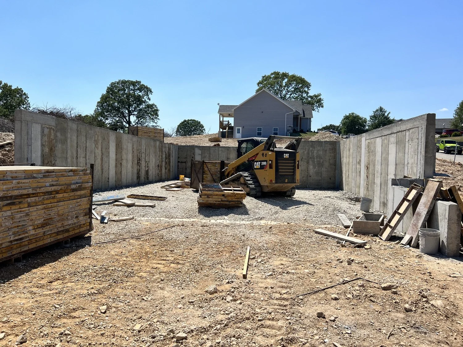 Construction site with concrete walls, a small excavator, construction materials, and a house in the background under a clear blue sky.