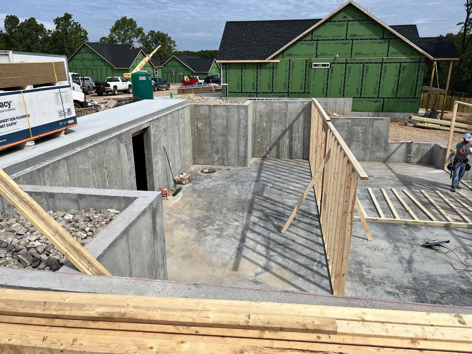 Construction site with partially built homes, showing concrete foundation walls, wooden framing, construction tools, and workers, with houses in green sheathing in the background.