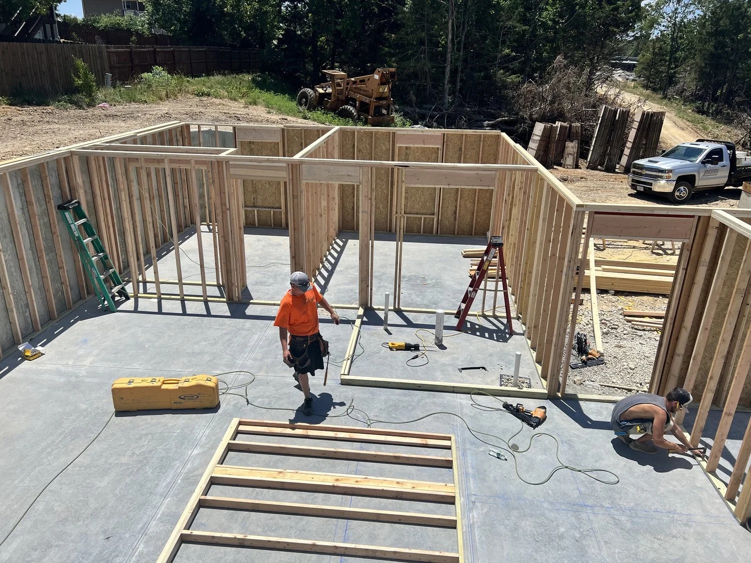 Construction workers building the wooden frame of a house on a concrete foundation, with tools and ladders visible, at a construction site surrounded by trees and a parked truck.
