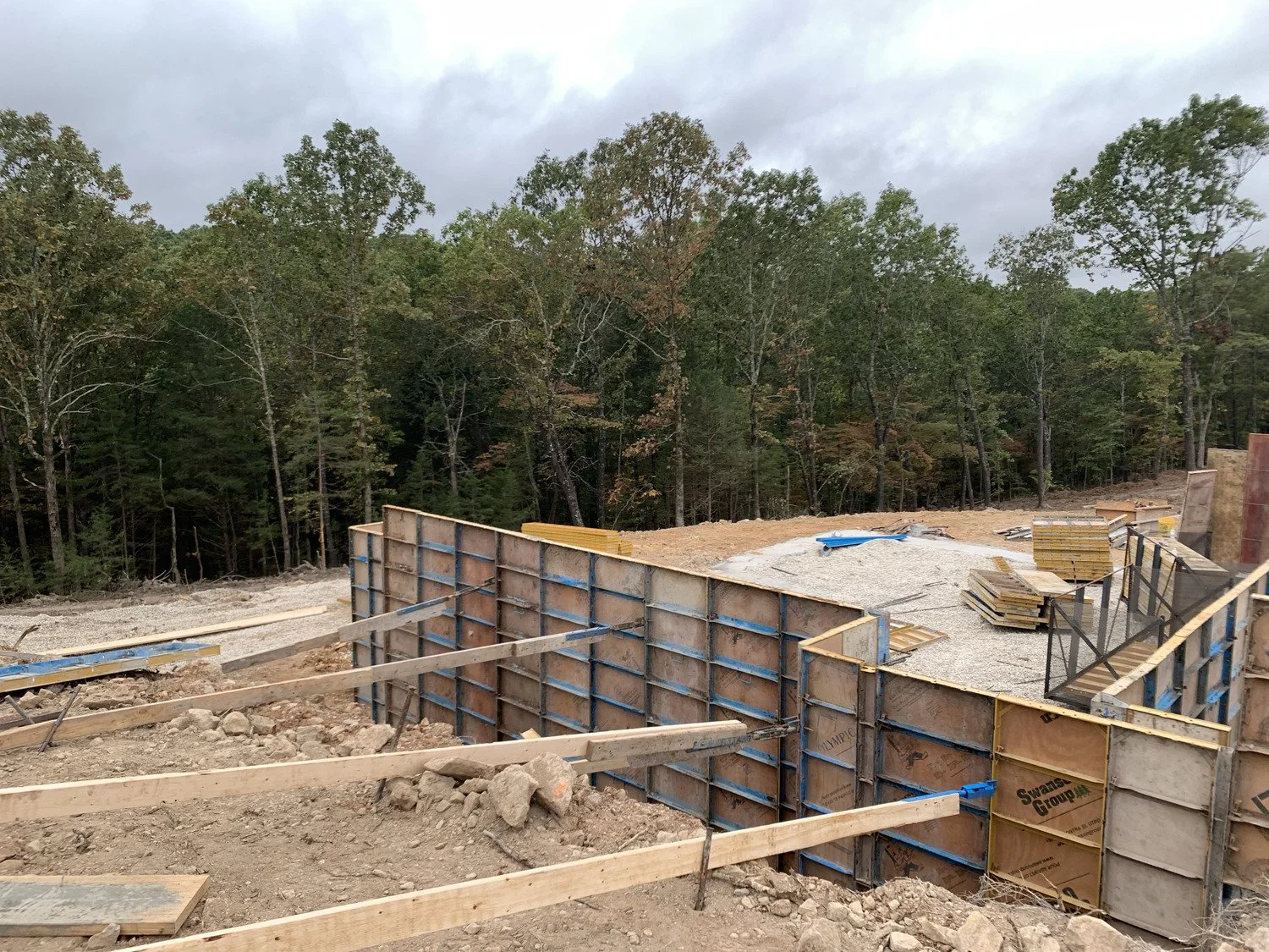 Construction site with wooden and metal framing for a wall, dirt ground, and a backdrop of trees and cloudy sky.