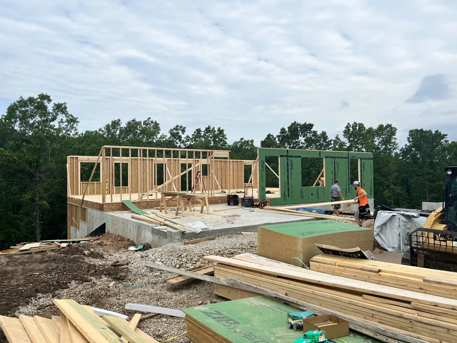Construction site with a house frame being built, workers installing green exterior sheathing, wood scattered around, and trees in the background during daytime.