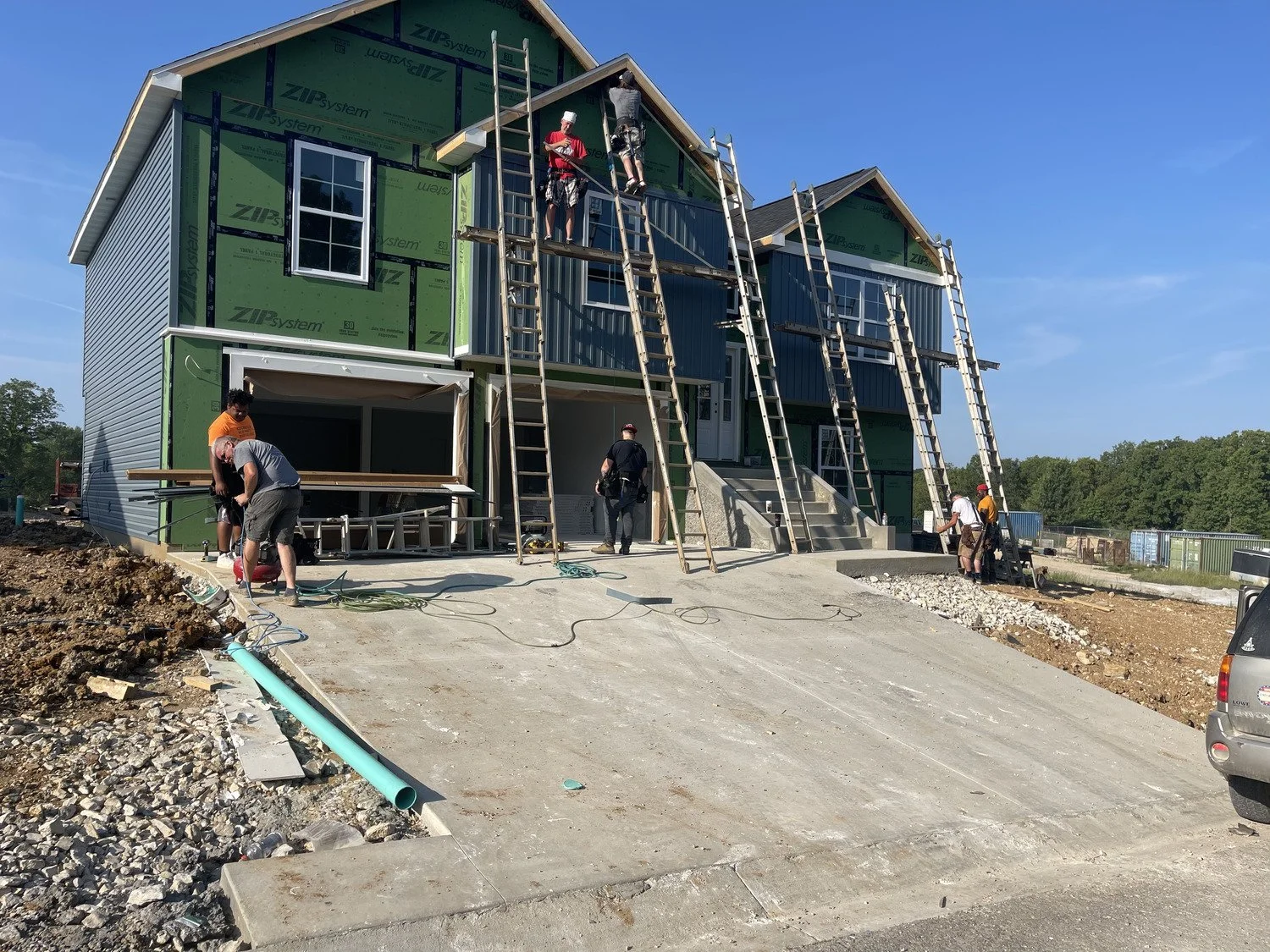 Several construction workers are building a multi-story house, with some working on the roof and others on the ground, surrounded by construction materials and equipment.