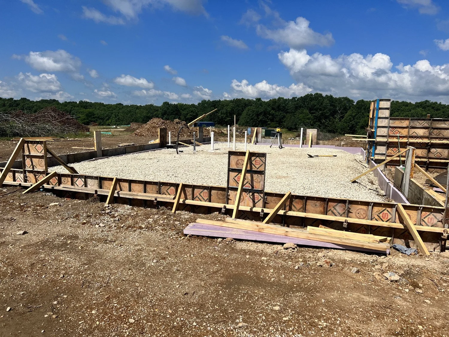 Construction site with a concrete foundation being prepared, surrounded by wooden framing and construction tools, under a partly cloudy sky.