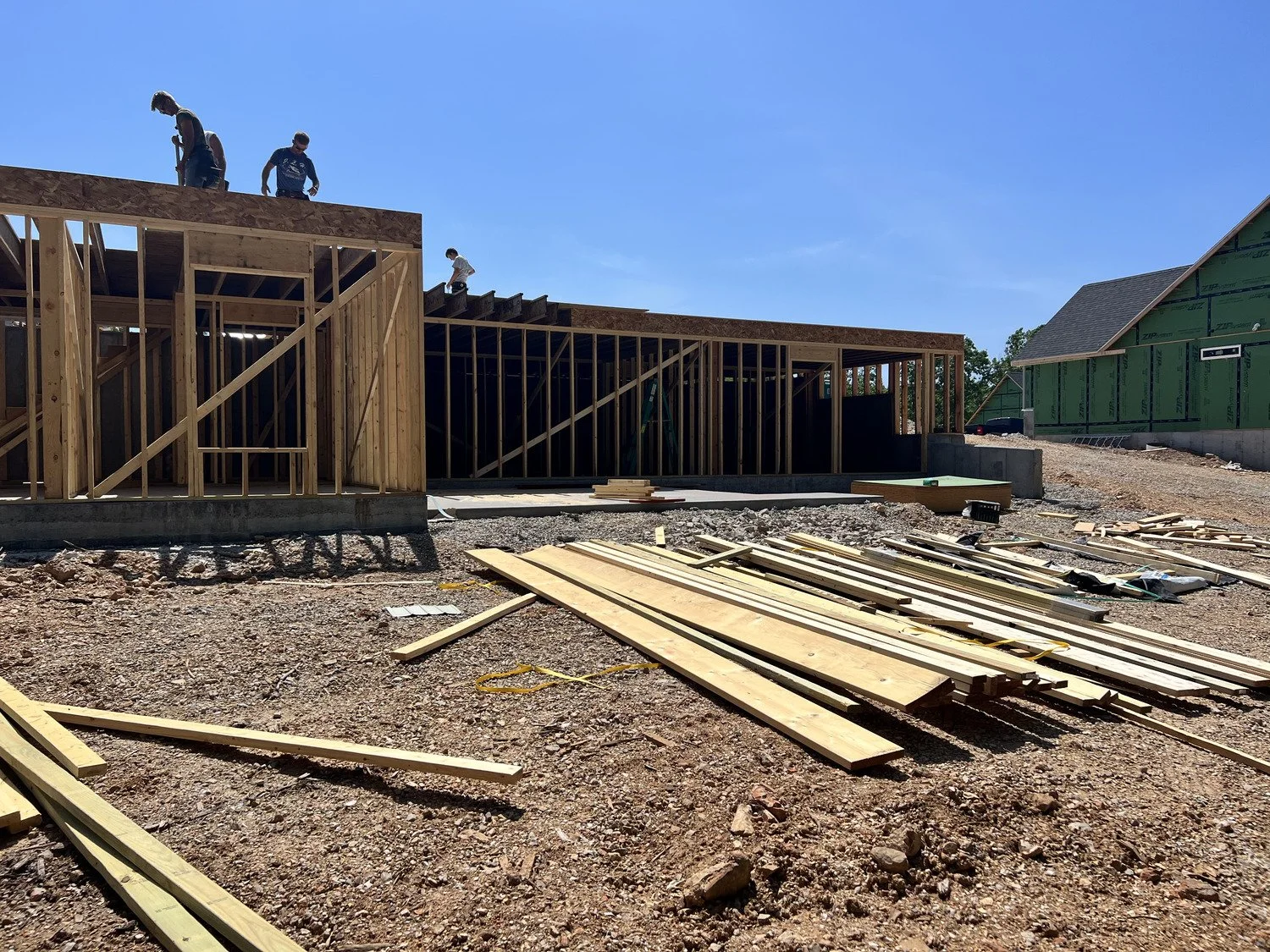 Construction site with wooden frames for a building, construction workers on the roof, and scattered lumber on the ground under a clear blue sky.