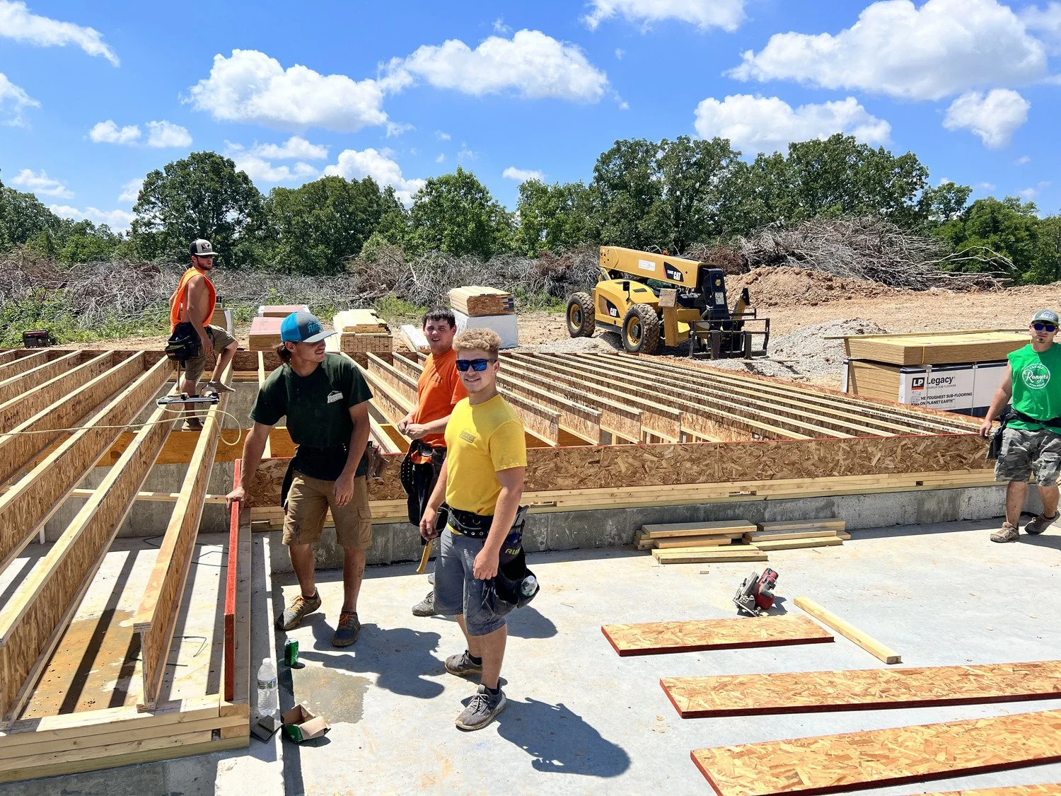 Construction workers assembling the wooden frame of a building on a sunny day. There are five workers, some smiling at the camera, with tools and construction materials around them. In the background, there is a yellow forklift and a pile of dirt and