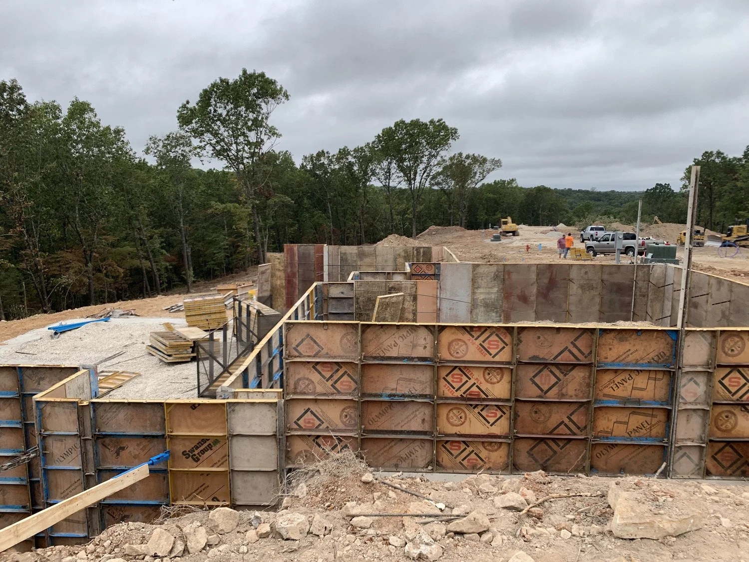 Construction site with concrete forms, construction workers, trucks, and construction equipment in a wooded area on cloudy day.
