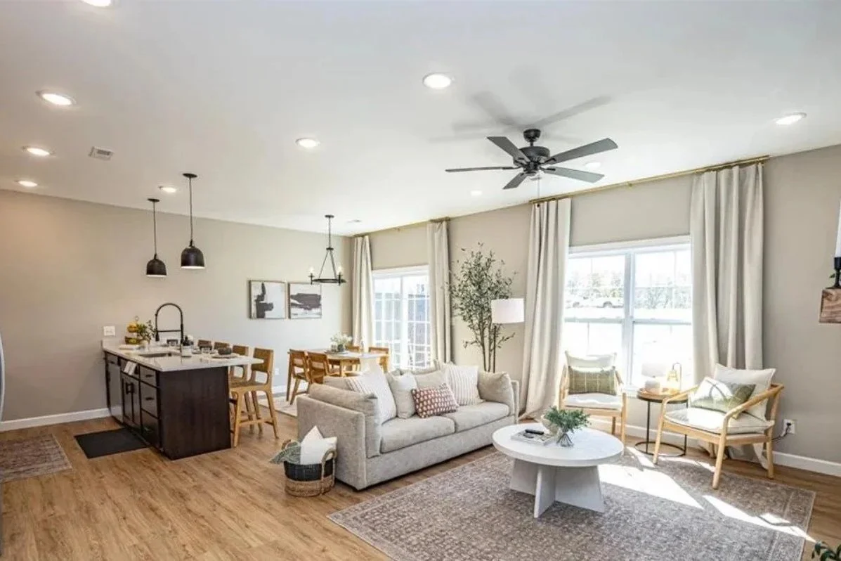 Living room with beige sofa, two armchairs, a round coffee table, large windows with curtains, ceiling fan, and a dining area with a table and chairs in the background.