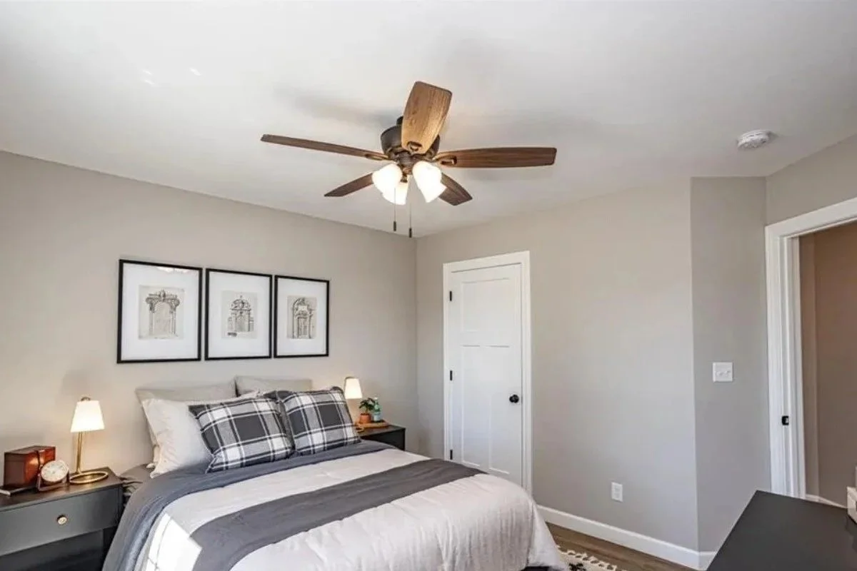 A bedroom with a bed, two bedside tables with lamps, framed pictures on the wall, a ceiling fan, and a door.