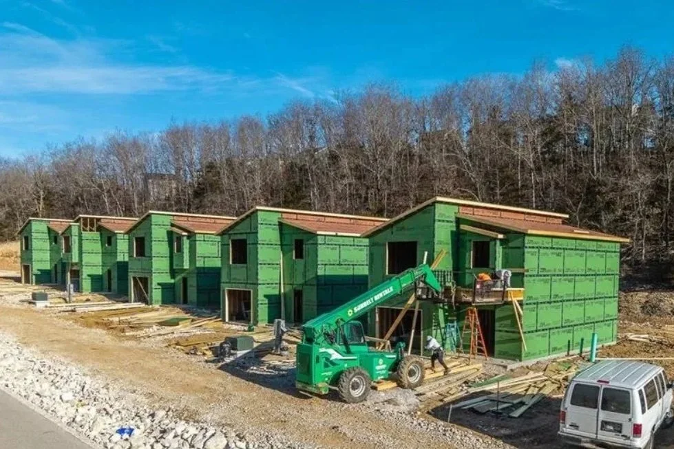 A row of green houses under construction with a crane, workers, and building materials outside.