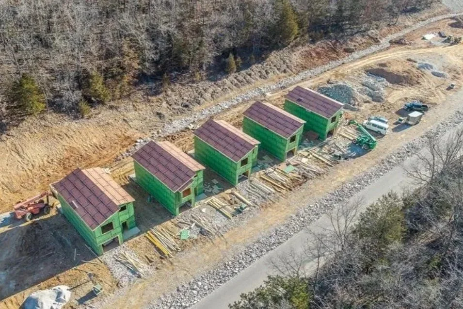Aerial view of five green houses under construction along a dirt road, with construction equipment and materials nearby, and a forested hillside in the background.