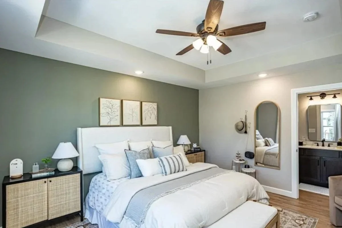A bedroom with a white bed, nightstands, lamps, a wall mirror, and a view into an en suite bathroom.