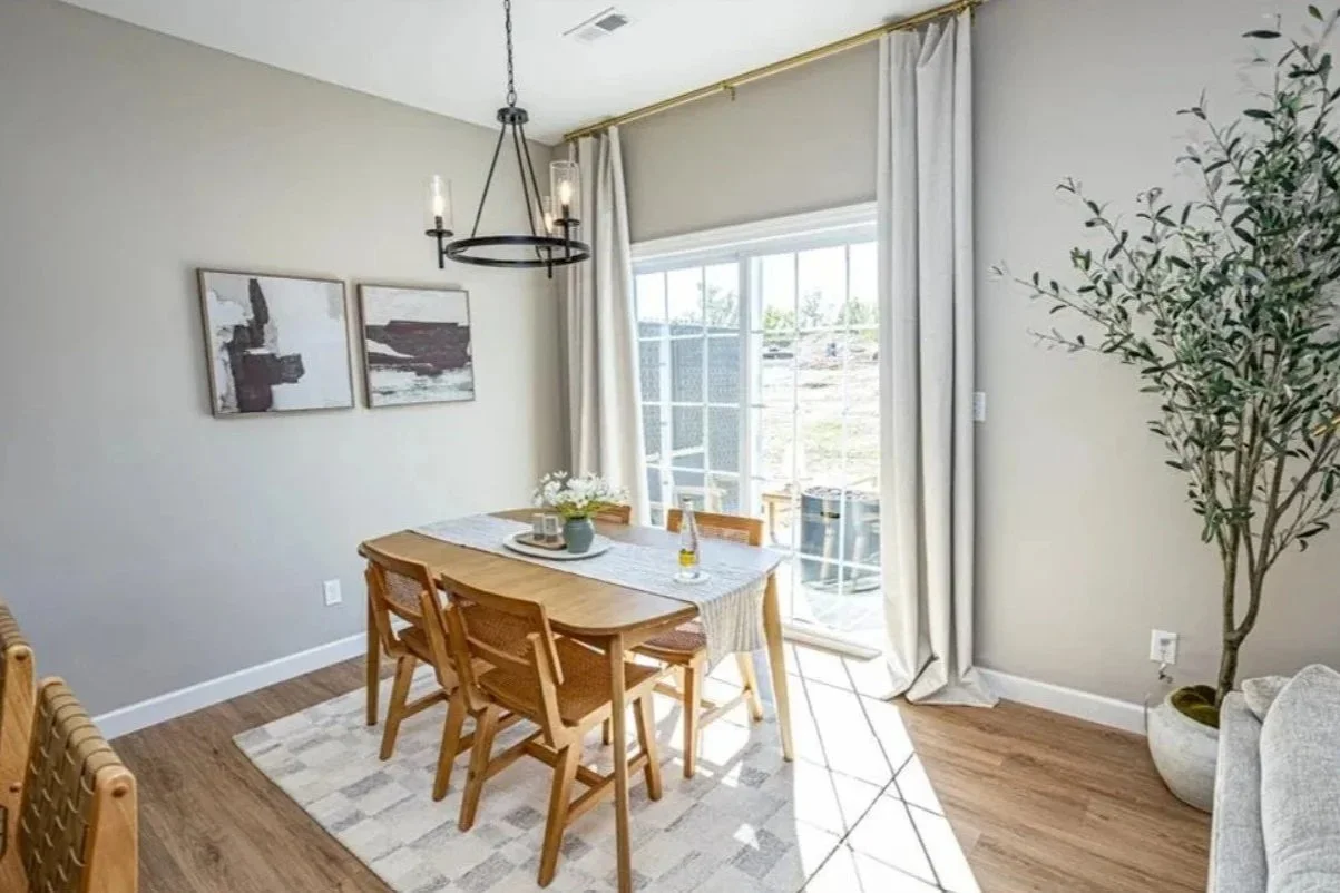 Dining room with wooden table, six chairs, floral centerpiece, glass sliding door with curtains, hanging chandelier, wall art, large potted plant, and hardwood floors.