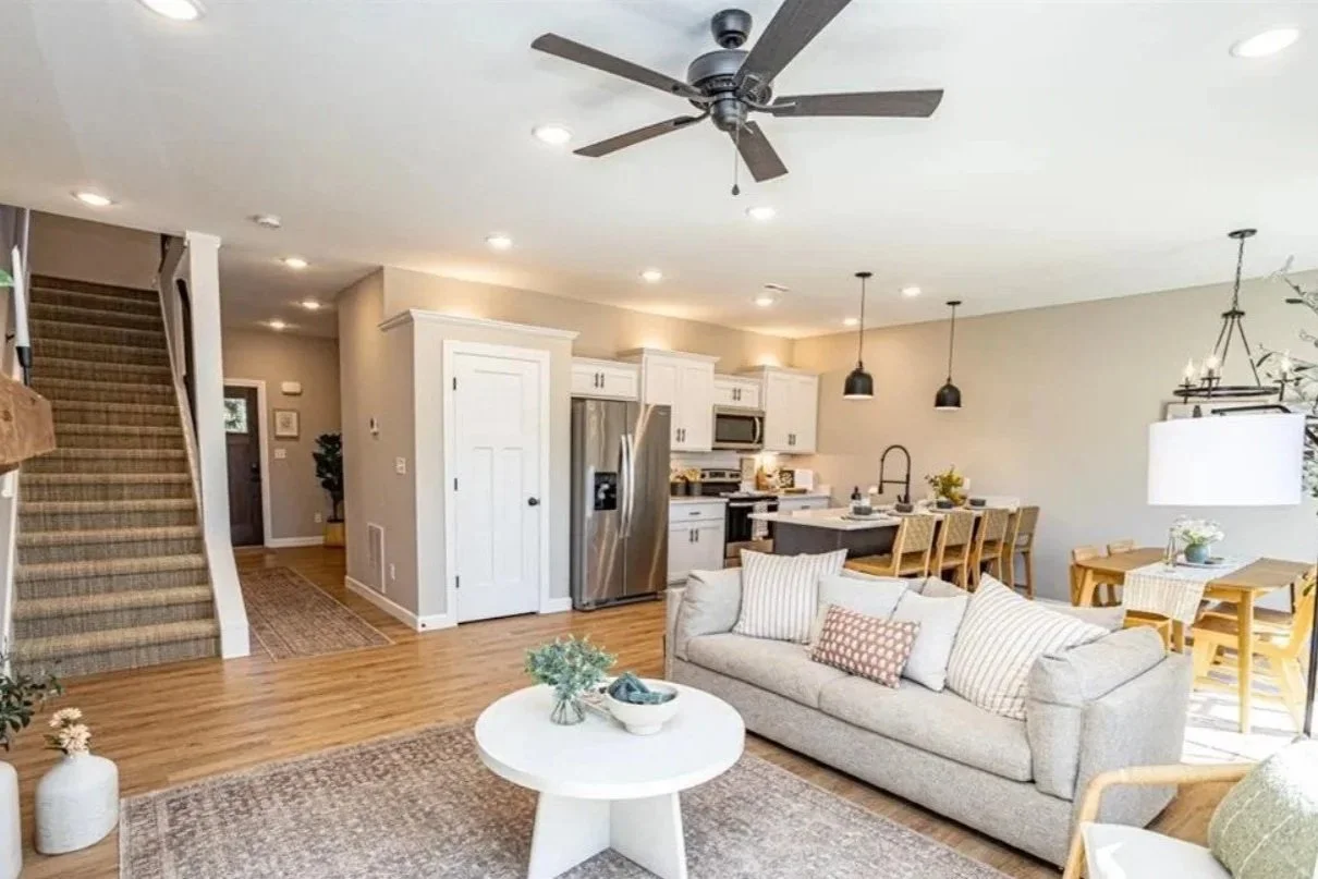 Open-concept living area with a beige sofa, round white coffee table decorated with a small vase and bowl, hardwood flooring, and a kitchen with white cabinets, stainless steel refrigerator, and a dining area with wooden table and chairs. Ceiling fan