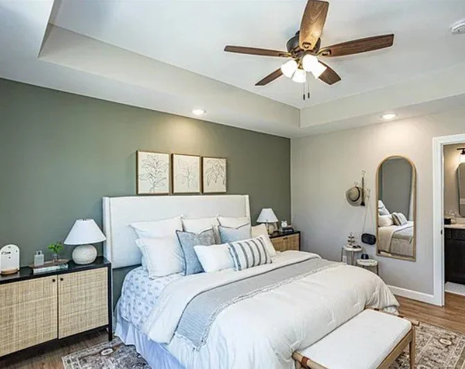 Bedroom with a white bed, green accent wall, three framed botanical art pieces, two white bedside lamps, a mirror, and a ceiling fan with wooden blades.