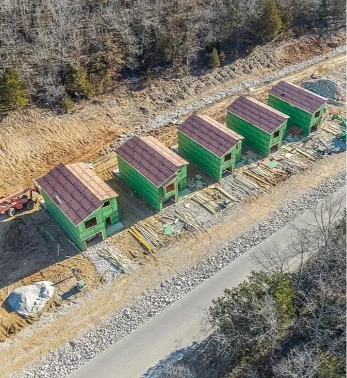Five small green houses under construction along a dirt road with construction materials nearby, surrounded by trees and a rocky hillside.