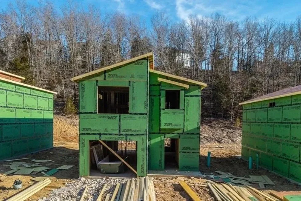 Three green framed houses under construction on a hillside with trees in the background and construction materials on the ground.