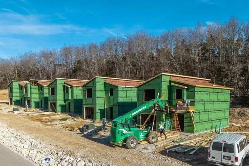 Multiple green unfinished houses under construction with a crane and workers, on a cleared construction site near a road.