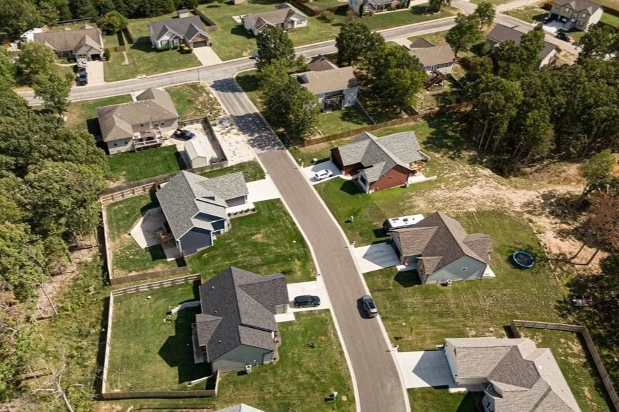 Aerial view of a suburban neighborhood showing houses, lawns, trees, driveways, and parked cars along a curved street.