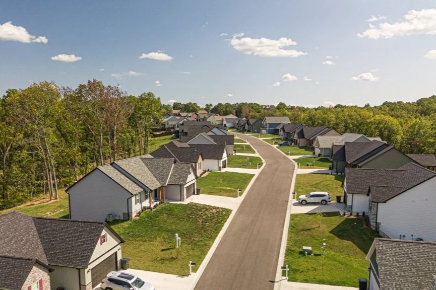 A suburban neighborhood street with new houses on both sides, some with cars parked in driveways, green lawns, and trees in the background under a partly cloudy sky.