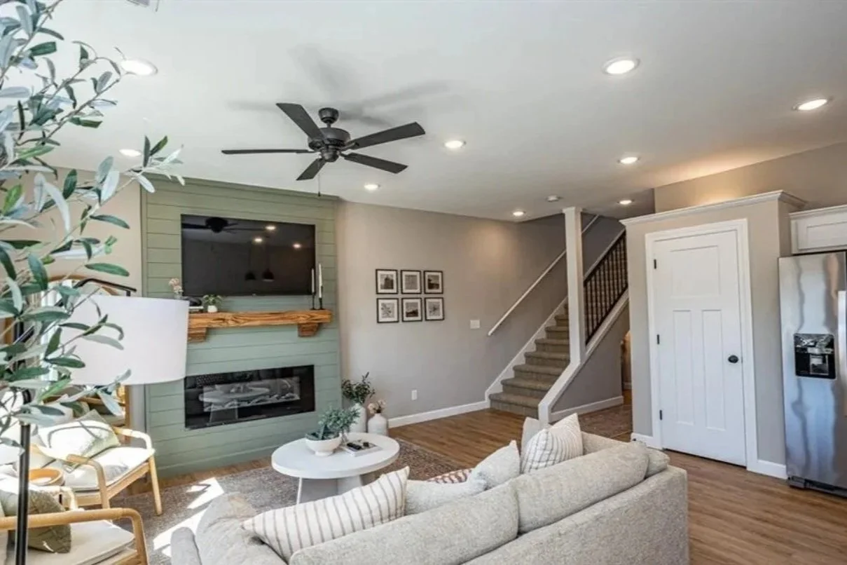 Living room with a beige sofa, wooden chairs with white cushions, a white coffee table with decorative items, a dark green accent wall with a mounted TV, a fireplace below the TV, a staircase with carpeted steps, a ceiling fan, and a stainless steel 