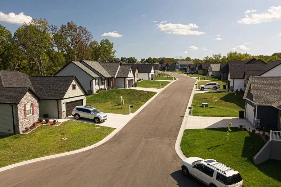 A quiet suburban neighborhood with new single-family houses, green lawns, and parked cars along a curved street during daytime.