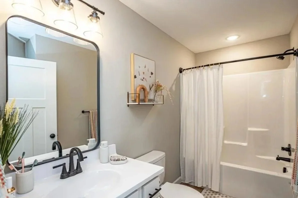 A modern bathroom with a white vanity, black fixtures, large mirror, potted plant, decorative items, white toilet, and a shower with a white curtain.