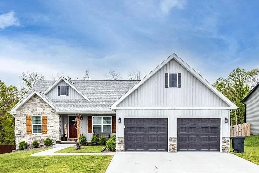 Front view of a suburban house with a two-car garage, white siding, stone accents, a small front porch, green lawn, and a blue sky.