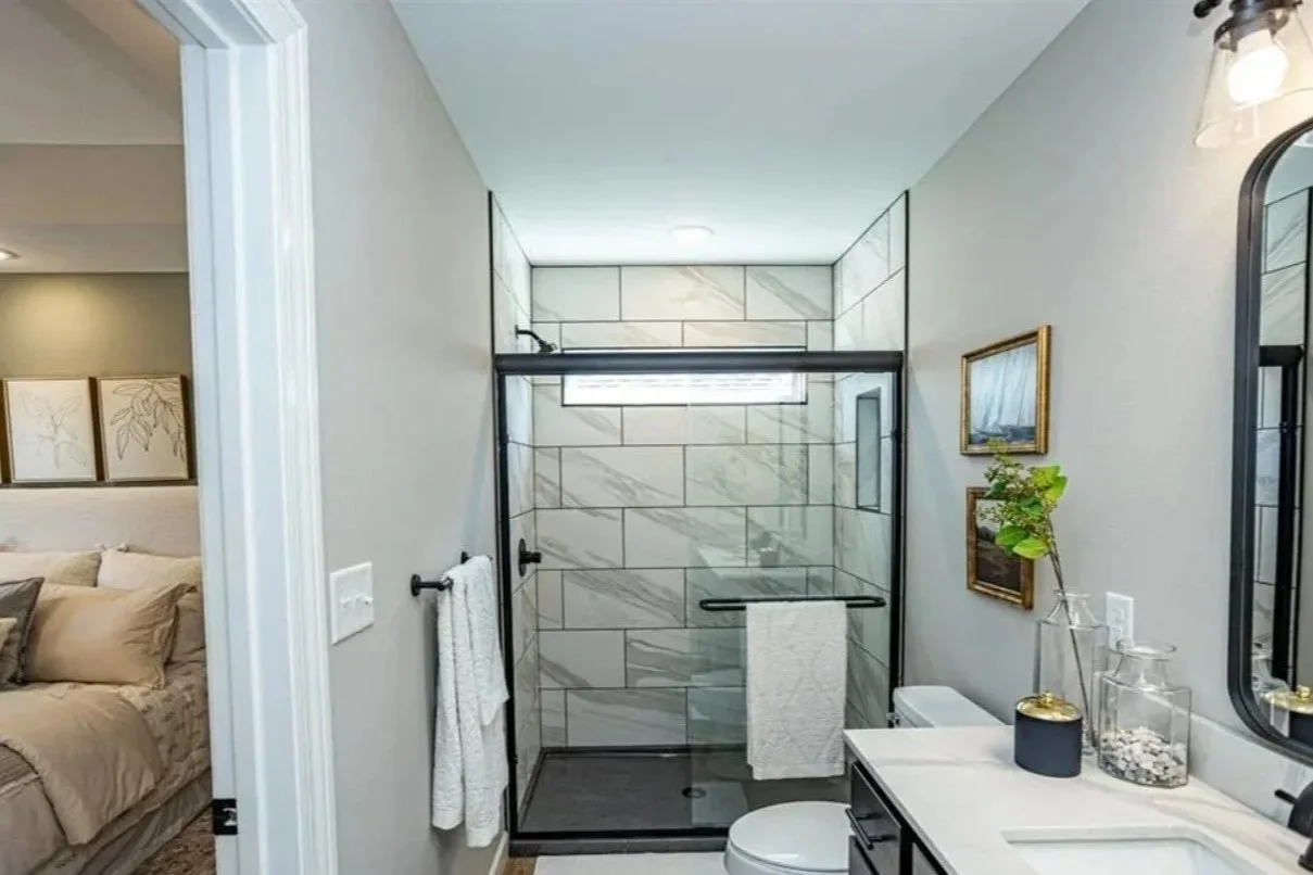 Modern bathroom with a shower enclosure, a white toilet, a vanity with a white countertop, a black faucet, and wall-mounted light fixtures. Decor includes framed artwork and decorative jars with stones and a plant.
