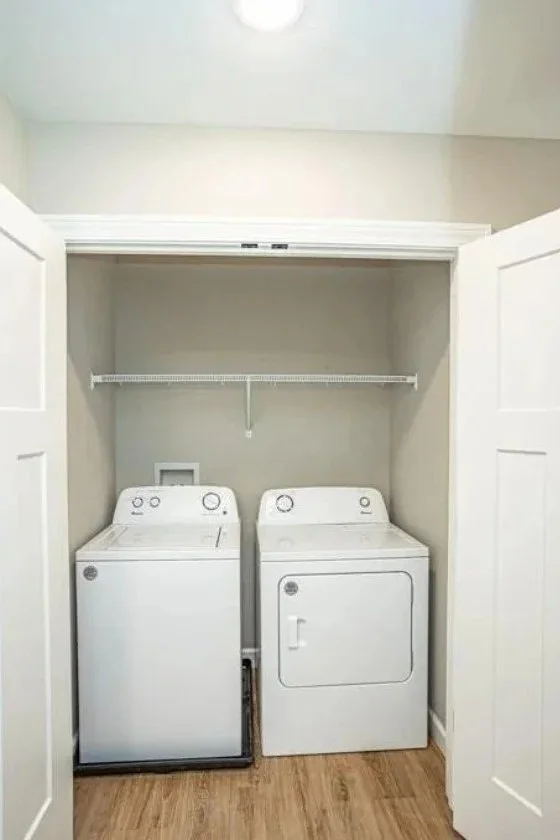 A laundry closet containing a white top-load washing machine on the left and a white dryer on the right, inside a small, beige-colored space with a wire shelf above.