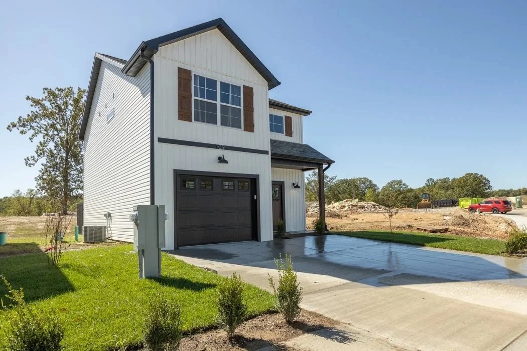 A modern two-story house with white siding and brown shutters, featuring a black garage door, a small front porch, and a newly paved driveway. The house is situated in a suburban area with trees and a clear blue sky.