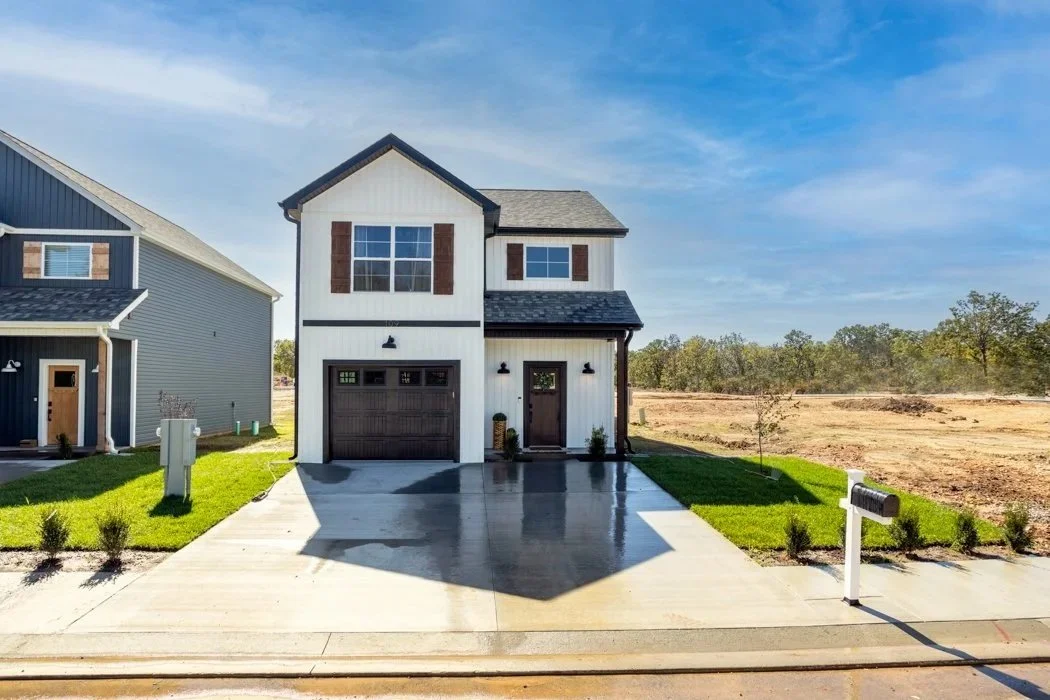 New two-story house with white exterior, dark brown garage door, and front door, with a concrete driveway and small front yard with grass and small bushes, on a clear day with blue sky and some nearby trees