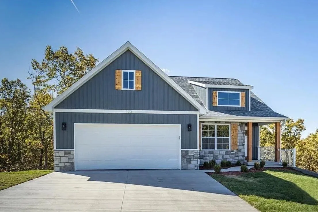 New blue house with gray stone accents, white garage door, and small front porch, surrounded by green lawn and trees.
