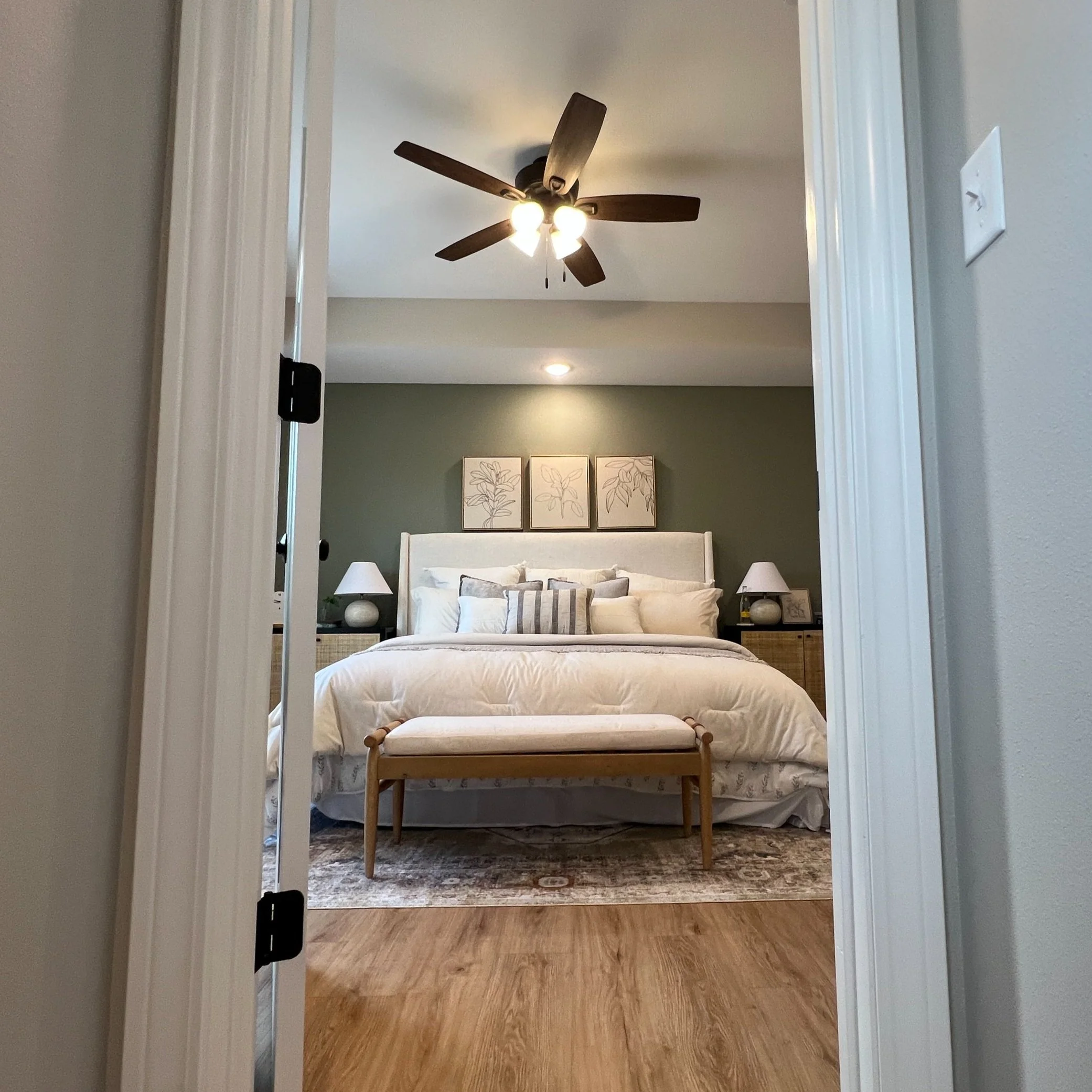 A bedroom viewed from a doorway featuring a white bed with multiple pillows against a green accent wall, framed botanical artwork above, two bedside tables with lamps, a ceiling fan, and wooden flooring with an area rug.