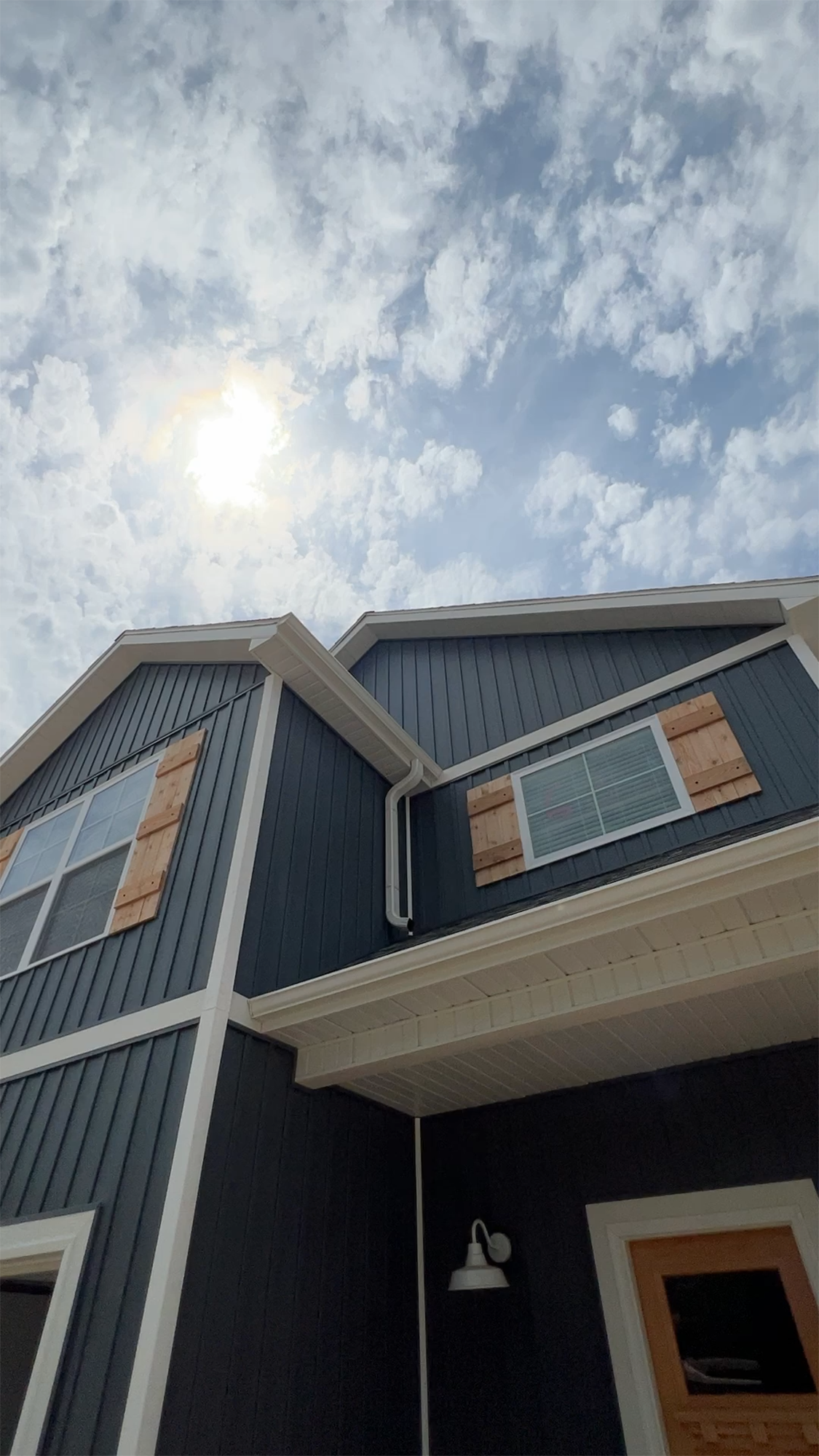 Upward view of blue house with white trim and honey-colored wooden accents, under a cloudy sky with sun.